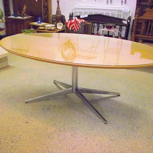 May include: Oval-shaped table with a light-colored, glossy top and a silver-colored, star-shaped base. The table's top has a light wood-colored border. The table is approximately 60 inches long and 30 inches wide.