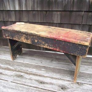 May include: A rustic wooden bench with a black painted finish and a worn, distressed look. The bench has a simple design with two legs and a flat top. The wood has a natural grain pattern and shows signs of age and use.