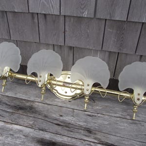 May include: A four-light vanity fixture featuring a polished brass horizontal bar and a central mounting plate. Each light is adorned with a frosted glass shade shaped like a scalloped shell, held in place by brass hardware and curved arms.