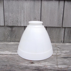 May include: A white, opaque glass lampshade with a textured, horizontal ribbed pattern. The shade has a flared, conical shape with a rounded top and a flat base. It sits on a weathered wooden surface against a gray wooden background.