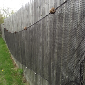 May include: A black mesh net fence with wooden floats attached, draped over a weathered gray wooden fence. The net is secured with small metal fasteners along the bottom edge. Green grass is visible at the base of the fence.