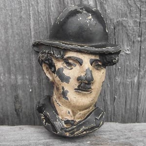 May include: A vintage bust of a man wearing a bowler hat. The sculpture has a black hat and coat, with a beige face and peeling paint. The bust is mounted on a weathered wooden surface.
