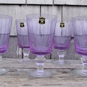 May include: A set of six vintage purple-tinted stemmed glasses. Each glass features a bulbous base, a clear stem, and a wide, slightly flared bowl. A gold and black emblem is affixed to each glass. The glasses are arranged on a weathered wooden surface.