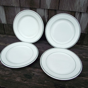 May include: Four white ceramic plates with a green trim. The plates are round and have a simple, elegant design. They are arranged on a dark wooden surface, creating a classic and clean aesthetic. Ideal for serving meals or as decorative pieces.
