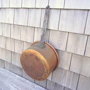 May include: A vintage copper pot with a long, metal handle. The pot has a worn, aged patina and is hanging on a wooden wall.
