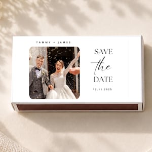 May include: A rectangular matchbox featuring a white label with a photograph of a couple in wedding attire. The text reads TAMMY + JAMES, SAVE the DATE, and 12.11.2025. The matchbox has a dark brown base and is displayed on a light surface.