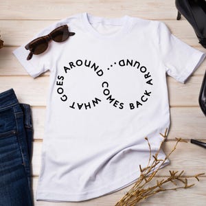 May include: White t-shirt with the phrase "WHAT GOES AROUND COMES BACK" in a circular design. The shirt is paired with sunglasses, dark blue jeans, and black high heels. A gold decorative item and sprigs of dried plants are also present.