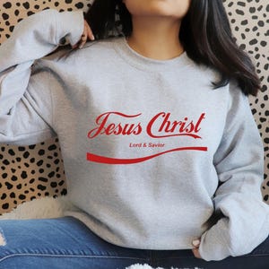 May include: A pale grey sweatshirt featuring the red text "Jesus Christ" in a script font, with "Lord & Savior" underneath. The design is similar to a well-known soft drink logo. The sweatshirt is worn with blue jeans.