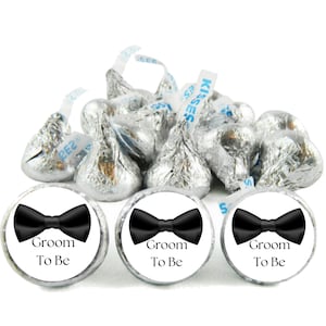 May include: A collection of foil-wrapped chocolate candies, some with the word "Kisses" printed on the paper strips. Three candies have white labels with a black bow tie graphic and the words "Groom To Be".