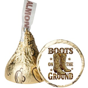 May include: A gold foil-wrapped chocolate candy with an almond inside, next to a white circular sticker. The sticker features a pair of gold cowboy boots with the text "BOOTS ON THE GROUND" in brown.