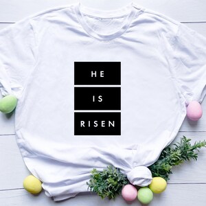 May include: White t-shirt featuring the text "HE IS RISEN" in a black rectangular design. The image includes pastel-colored Easter eggs and greenery, suggesting a seasonal or religious theme.