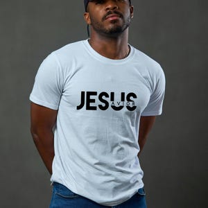 May include: A white t-shirt featuring the word "JESUS" above "SAVIOR" in black. The short-sleeved shirt is worn by a person. The person is wearing a black baseball cap and blue jeans.