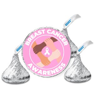 May include: Three Hershey's Kisses candies with silver foil wrappers. A pink circular emblem in the center reads "Breast Cancer Awareness" with a graphic of joined hands and a pink ribbon. The candy's paper flags read "Kisses".