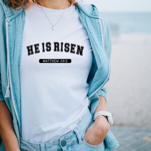 May include: White t-shirt with the text "HE IS RISEN" in black, arched lettering above "MATTHEW 28:6" in a black rectangle. The shirt is worn with a light blue denim jacket and jeans. A silver necklace and white watch are also visible.