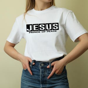 May include: White t-shirt featuring the word "JESUS" in black block letters, with "PRINCE OF PEACE" underneath. The top is short-sleeved and made of a soft fabric. The model is wearing blue denim jeans.