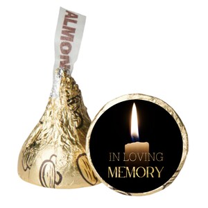 May include: A gold foil-wrapped Hershey's Kiss candy with an almond label, next to a black circle with a lit candle and the words "IN LOVING MEMORY" in gold lettering. The image is set against a white background.