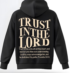 Könnte beinhalten: Schwarzer Kapuzenpullover mit dem Text "TRUST IN THE LORD" in cremefarbener, stilisierter Schrift. Auf der Rückseite des Kapuzenpullovers befindet sich auch ein Zitat aus Sprüche 3:5-6. Der Kapuzenpullover hat eine Kapuze und lange Ärmel.