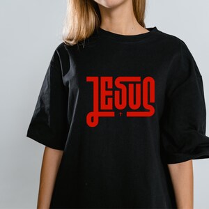 May include: Black t-shirt featuring the word "JESUS" in red, stylized lettering. The text is centered on the shirt, with a small cross symbol below the "U". The shirt has short sleeves and a relaxed fit.