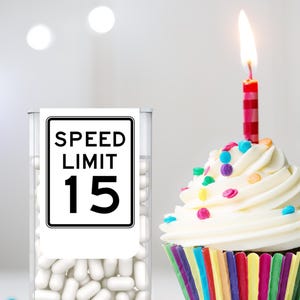 May include: A clear rectangular container filled with white pill-shaped candies, featuring a white label with a black border and the text SPEED LIMIT 15. Beside it sits a cupcake with white frosting, colorful sprinkles, and a lit red and pink striped candle.