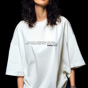 Puede incluir: Una camiseta blanca con el texto "BE NOT OVERCOME OF EVIL, BUT OVERCOME EVIL WITH GOOD ROMANS 12:21" impreso en negro. La camiseta tiene mangas cortas y cuello redondo.
