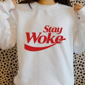 May include: White sweatshirt with the red text "Stay Woke" in a stylized font. The design is reminiscent of a popular soda brand's logo. The sweatshirt is a casual, comfortable style.