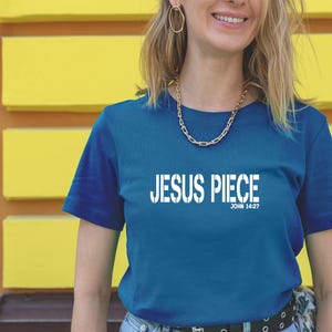May include: A teal short-sleeved t-shirt with the white text "JESUS PIECE" and "JOHN 14:27" underneath. The person wearing the shirt has blonde hair and is wearing gold jewellery.