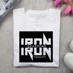 May include: White t-shirt with a black square graphic. The graphic reads "IRON SHARPENS IRON" in bold white letters, with "Proverbs 27:17" below. The shirt is displayed with white sneakers and blue jeans.