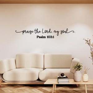 May include: A black wall decal featuring the cursive script text 'praise the Lord, my soul' followed by the text 'Psalm 103:1' in a bold, sans-serif font. The decal is applied to a plain, light-colored wall above a beige sofa and a wooden coffee table.
