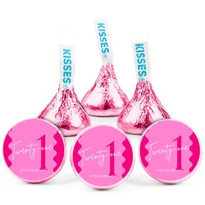 May include: Pink foil-wrapped chocolate candies and round stickers. The candies have white paper flags that say "Kisses". The stickers say "Twenty-one" with a large number "1" and "Let's Celebrate".