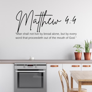 Op de afbeelding: Een keuken met witte kasten, een roestvrijstalen oven en een houten tafel. Op de muur staat de tekst "Matteüs 4:4" en een Bijbelcitaat. Twee potplanten en een vaas bloemen zorgen voor kleur.