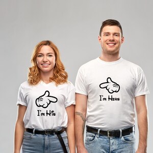 May include: Two people wearing white t-shirts with matching black cartoon hand designs. One shirt says "I'm His" and the other says "I'm Hers". Both shirts have a black belt and blue jeans.