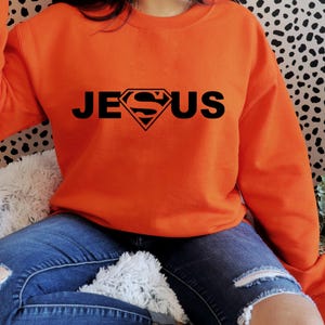 May include: An orange crew neck sweatshirt featuring the word "JESUS" in black, with a diamond-shaped logo replacing the "S". The sweatshirt is paired with blue denim jeans.