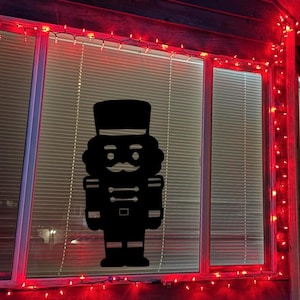 May include: A black silhouette of a nutcracker soldier is displayed in a window. The window is framed with red lights, and the blinds are partially closed. The nutcracker has a hat, mustache, and uniform.