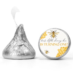 Puede incluir: Un caramelo de chocolate envuelto en papel de aluminio plateado junto a una pegatina redonda. La pegatina tiene un diseño de abeja y panal con el texto "Our little honey bee IS TURNING ONE!" en una fuente lúdica. El tema es un cumpleaños con temática de abejas.