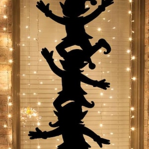 Puede incluir: Silueta negra de tres elfos apilados uno encima del otro, con los brazos y las piernas extendidos. Los elfos están frente a una ventana con persianas y luces de cadena, creando una escena festiva navideña.