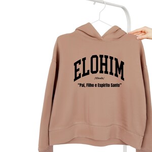 May include: A tan hoodie with the word "ELOHIM" in large black block letters across the chest. Below the word "ELOHIM" is the text "/Eloah/" and the phrase "Pal, Filho e Espirito Santo" in black. The hoodie is hanging on a white hanger.