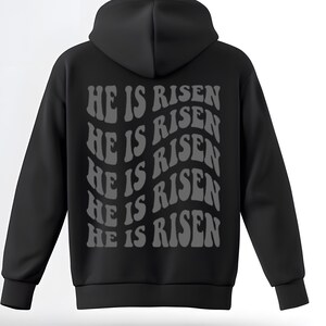 May include: Black hooded sweatshirt with a drawstring and long sleeves. The back displays the phrase "HE IS RISEN" in a wavy, grey font. The sweatshirt is made of a soft material.