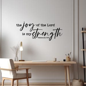 Pode incluir: Um adesivo de parede preto com o texto "the joy of the Lord is my strength" em uma combinação de fontes serifadas e manuscritas, com a citação "Nehemiah 8:10" posicionada abaixo da palavra "strength" em uma fonte sem serifa menor.