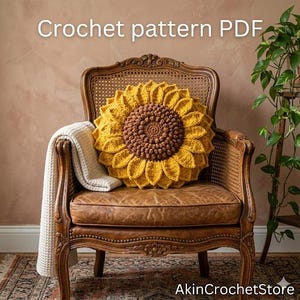 Op de afbeelding: Een gehaakt zonnebloemkussen met een bruin midden en gele bloemblaadjes ligt op een vintage leren stoel. Een cr&egrave;mekleurige deken is over de arm van de stoel gedrapeerd. De tekst "Crochet pattern PDF" is zichtbaar.