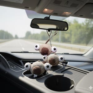 May include: A collection of crocheted car accessories, each a round creature with large, googly eyes. The creatures are in shades of brown and beige, with white and black eye details. One hangs from the rearview mirror, others sit on the dashboard.