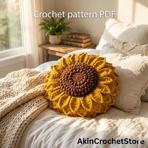 Op de afbeelding: Een gehaakt zonnebloemkussen met een bruin midden en gele bloemblaadjes ligt op een bed met wit linnen. De tekst "Crochet pattern PDF" is bovenaan zichtbaar en "AkinCrochetStore" onderaan. Een cr&egrave;mekleurige deken is op het bed gedrapeerd.