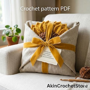 Op de afbeelding: Een gehaakt zonnebloemkussen verpakt in een linnen stof, vastgebonden met een mosterdgeel fluwelen lint. Het kussen ligt op een neutraal gekleurde fauteuil. De tekst "Crochet pattern PDF" en "AkinCrochetStore" zijn zichtbaar.