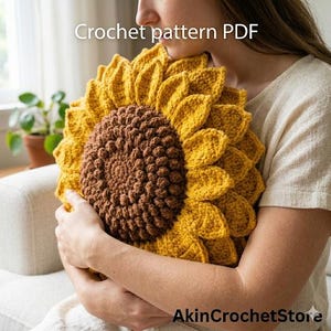 Op de afbeelding: Een gehaakt zonnebloemkussen met een bruin midden en gele bloemblaadjes wordt vastgehouden. Het kussen is handgemaakt en heeft de tekst "Crochet pattern PDF" en "AkinCrochetStore".