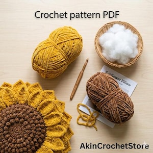 Op de afbeelding: Een gehaakt zonnebloemkussen met gele en bruine wol, een houten haaknaald en een mandje met witte vulling. De afbeelding bevat ook twee strengen garen en een gedrukt patroon met de tekst "Sunflower Pillow". De tekst "Crochet pattern PDF" staat bovenaan.