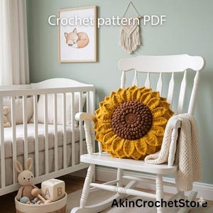 Op de afbeelding: Een gehaakt zonnebloemkussen in geel en bruin ligt op een witte schommelstoel. Een cr&egrave;mekleurige deken is over de arm van de stoel gedrapeerd. De tekst "Crochet pattern PDF" is zichtbaar in de afbeelding.