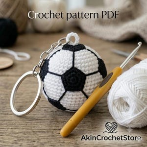 May include: A crochet pattern PDF for a soccer ball keychain. The image shows a small, spherical, black and white crocheted soccer ball attached to a silver metal keyring, alongside a yellow crochet hook and a ball of white yarn.