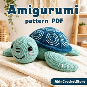 May include: A crochet amigurumi turtle with a light green head and body, featuring a dark blue shell with white stitched hexagonal patterns. The turtle has black safety eyes, a stitched smile, and pink cheeks. Text reads Amigurumi pattern PDF.
