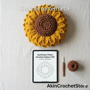 Op de afbeelding: Een gehaakt zonnebloemkussen met een bruin midden en gele bloemblaadjes. Een tablet toont een haakpatroon voor een zonnebloemkussen. Een haaknaald en een bol bruin garen zijn ook aanwezig. De tekst "Crochet pattern PDF" en "Sunflower Pillow Crochet Pattern PDF" zijn zichtbaar.