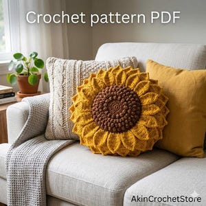 Op de afbeelding: Een gehaakt zonnebloemkussen met een bruin midden en gele bloemblaadjes ligt op een cr&egrave;mekleurige bank, naast een kabelgebreid kussen en een mosterdgeel kussen. De tekst "Crochet pattern PDF" is zichtbaar.