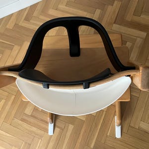 May include: A modern, minimalist rocking chair with a black backrest and seat, and a white, curved base. The chair has wooden side supports and is designed for comfort and style. The chair is set on a wooden surface.